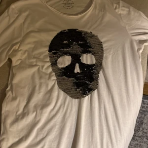 Vit t-shirt med paljett-skalle Jack & Jones - Säljer en vit t-shirt från Jack & Jones Originals med korta ärmar. På framsidan finns en stor svart skalle gjord av paljetter som ger en cool och edgy vibe. Materialet är mjuk bomull och passformen är normal. Perfekt för dig som gillar statement-plagg.