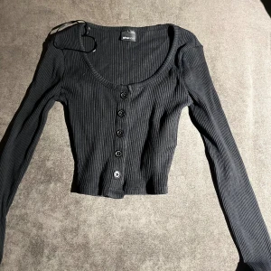 Svart ribbad croppad topp Gina Tricot - Säljer en svart, croppad topp från Gina Tricot i storlek XXS. Toppen har ribbad struktur, lång ärm och knappar framtill. Den har en rundad halsringning och är figurnära i passformen. Perfekt för dig som gillar stilrena och enkla plagg med snygga detaljer.