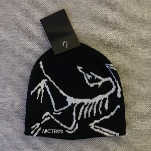 Svart mössa arc teryx med vit grafik - Svart mössa från Arc'teryx med stor vit grafisk dinosaurie på framsidan och logga. Snygg, stilren design som sticker ut och passar perfekt för dig som gillar outdoor eller streetstyle. Materialet är stickat och känns mjukt mot huvudet.