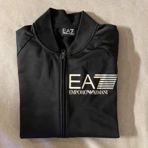 EA7 Emporio Armani kofta - Svart bomberjacka från EA7 Emporio Armani med stor vit logga på bröstet. Jackan har dragkedja framtill, ribbade muddar och klassisk krage. Tillverkad i mjuk polyester för en sportig och clean look. Använd men mycket väl omhändertagen. Först till kvarn!