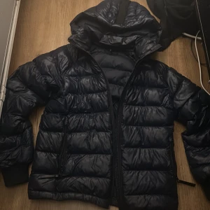 Mörkblå Peak Performance dunjacka - Säljer en mörkblå dunjacka från Peak Performance med huva och snygga detaljer som logotyp på ärmen och huvan. Jackan har quiltad design, dragkedja framtill och ribbade muddar vid ärmslut. Perfekt för kalla dagar och har en modern, loose passform.