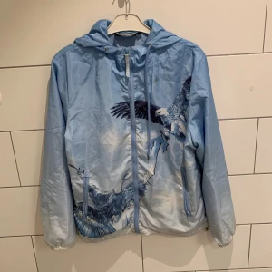 Trapstar vindjacka - Säljer en blå vindjacka från Trapstar med snyggt örntryck på både fram- och baksida. Jackan har huva med dragsko, dragkedja framtill och två fickor. Lätt material som passar perfekt för streetwear-stil. Trapstar-logga broderad på ryggen. Storlek M men passar som S. Skada vid armhåla (se bild 5)