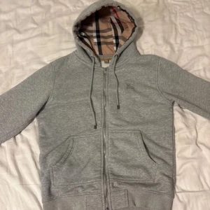 Grå Burberry hoodie med dragkedja - Snygg grå hoodie från Burberry med klassiskt rutigt foder i huvan och broderad logga på bröstet. Tröjan har dragkedja, snörning i huvan och två stora fickor framtill. Tillverkad i mjuk bomull med lite stretch för extra komfort. Kan gå ner i pris vid snabb affär 