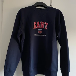 Mörkblå sweatshirt från GANT XS - Mörkblå sweatshirt från GANT med röd och vit logga framtill. Klassisk rund halsringning, ribbade muddar och lång ärm. Skön och stilren tröja i bomull, perfekt för en avslappnad look.