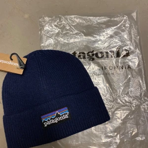 Mörkblå ribbad mössa från Patagonia - Snygg mörkblå mössa från Patagonia med ribbad struktur och klassisk logopatch framtill i färgglada toner. Mössan har uppvikt kant och är tillverkad i mjukt stickat material som håller dig varm under kyliga dagar.