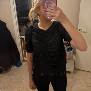 Svart puffärmad blus från H&M - Svart blus från H&M med korta puffärmar och snygg rynkdetalj framtill. V-ringad hals och lätt glansigt material som ger en cool look. Perfekt till jeans eller kjol för en trendig outfit.