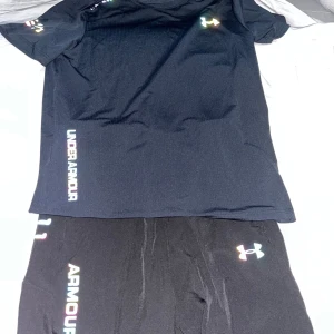 Svart Under Armour tracksuit i funktionsmaterial - Svart tracksuit från Under Armour i lätt och snabbtorkande funktionsmaterial. Klassisk rund hals, korta ärmar och logga i regnbågsfärger på bröstet och längs sidan. Perfekt för dig som gillar sportig och clean stil.
