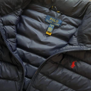 Mörkblå dunjacka från Polo Ralph Lauren - Snygg mörkblå dunjacka från Polo Ralph Lauren med röd broderad logga på bröstet. Jackan har quiltad design, hög krage, dragkedja framtill och två fickor med dragkedja. Perfekt för kyliga dagar och riktigt clean look. Använd fåtal gånger som ny. Junior storlek ej vuxen . ( L 14-16 år ) 