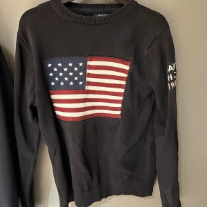 Svart stickad tröja GANT med USA-flagga - Svart stickad tröja från GANT med stor USA-flagga i rött, vitt och blått på bröstet. Broderad text på ena ärmen med 'GANT H-C 1949'. Rund hals och långa ärmar, perfekt för dig som gillar klassisk college-stil.
