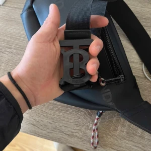 Svart axelväska från Burberry - Snygg svart axelväska från Burberry med tydlig logga och coolt TB-spänne i matt svart. Väskan har dragkedja framtill och justerbar rem med detaljer i textil och rep. Perfekt för att bära dina essentials med stil.
