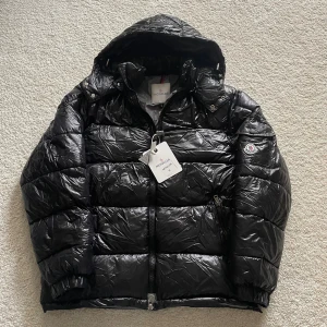Svart Moncler dunjacka med huva - Säljer en svart, glansig dunjacka från Moncler med huva och dragkedja. Jackan har puffig design, klassisk Moncler-logga på ärmen och snygga detaljer som tryckknappar vid kragen. Perfekt för kalla dagar och riktigt stilren vinterlook.