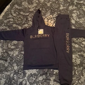 Mörkblå Burberry hoodie med broderi - Snygg mörkblå hoodie från Burberry med broderad logga i beige, rött och svart på bröstet. Tröjan har huva med snörning och en klassisk känguruficka framtill. Materialet är mjuk bomull och passformen är normal. Perfekt för dig som gillar exklusiva streetwear-vibes.