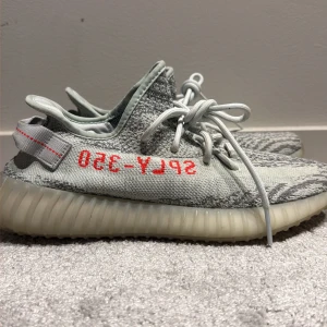 Adidas Yeezy Boost 350 V2 Blue Tint - Säljer ett par Adidas Yeezy Boost 350 V2 Blue Tint med grått och ljusblått stickat mönster, röd SPLY-350 text på sidan och genomskinlig ribbad sula. Skorna har snörning och är tillverkade i syntetmaterial med en bekväm boost-sula.