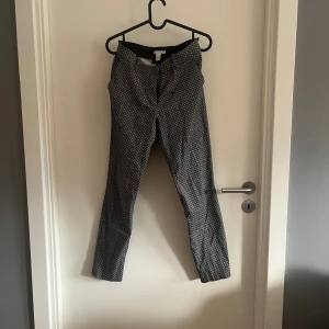 Svarta mönstrade byxor från H&M - Svarta byxor från H&M i storlek 36 med vitt rutmönster. De har en rak passform och midwaist, vilket ger en clean och modern look. 