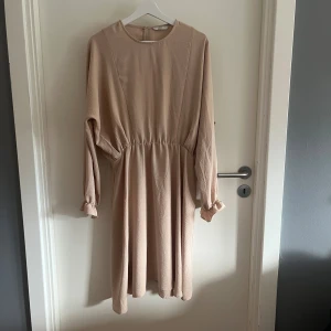 Beige långärmad klänning från ONLY - Beige klänning från ONLY med lång ärm och rund halsringning. Modellen har resår i midjan för en snygg siluett och diskreta volangdetaljer vid ärmsluten. 