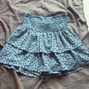 Blå volangkjol med blommigt mönster - Supersöt blå kortkjol med svarta små blommor och volanger. Kjolen har en bred, smockad midja som ger en bekväm och snygg passform. Perfekt för dig som gillar en feminin och lekfull stil. Materialet känns mjukt och lätt mot huden.