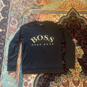 Svart BOSS tröja med guldtryck - Svart långärmad tröja från Hugo Boss i storlek S. Snyggt guldigt BOSS-tryck på bröstet och klassisk rund halsringning. Tröjan är tillverkad i mjuk bomull och har ribbade muddar vid ärmslut och nederkant. Perfekt för dig som gillar stilrena märkesplagg.
