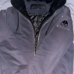 Svart bomberjacka Moose Knuckles - Snygg svart bomberjacka från Moose Knuckles med quiltat innerfoder och mjuk fuskpäls i luvan. Jackan har dragkedja, ribbade muddar och ikonisk logga på ärmen. Perfekt för kalla dagar och riktigt stilren look. Tags finns