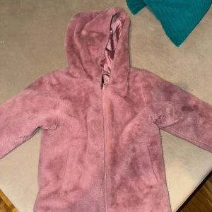 Rosa teddyjacka från Name It - Mjuk och fluffig rosa teddyjacka från Name It med huva och dragkedja framtill. Jackan har ett lent, glansigt foder och är perfekt för kyliga dagar. Enkel design utan mönster eller extra detaljer, passar dig som gillar mysiga och stilrena plagg.