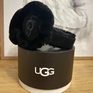 Svarta öronmuffar från UGG i päls - Mysiga svarta öronmuffar från UGG med broderad logga på sidan. Tillverkade i äkta fårskinnspäls och mocka för extra värme och lyxig känsla. Perfekta för kalla vinterdagar när du vill hålla stilen och värmen.
