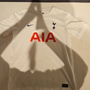 Tottenham Hotspur Nike fotbollströja XL - Tottenham Hotspur matchtröja från Nike i vitt med klubbmärke och Nike-logga på bröstet samt röd AIA-sponsor. Tillverkad i Dri-FIT-material som andas, kortärmad och med V-ringning. Perfekt för dig som vill visa ditt stöd på läktaren eller planen. Den har lite fläckar på magen som man kan se på första och sista bilden.