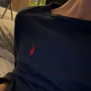 Mörkblå långärmad tröja Polo Ralph Lauren - Snygg mörkblå långärmad tröja från Polo Ralph Lauren med klassisk röd broderad logga på bröstet. Tröjan är i mjuk bomull och har en enkel, stilren design med rund hals. Perfekt för dig som gillar en clean och tidlös look.
