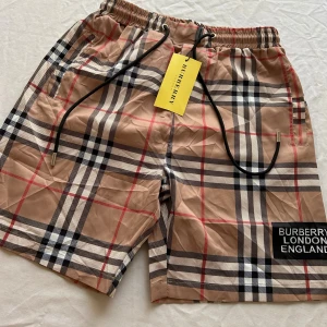Rutig Burberry shorts med snörning - Snygga shorts  Burberry med klassiskt rutigt mönster i beige, svart, vitt och rött. De har elastisk midja med svart snörning och Burberry London England-tryck på benet. Perfekta för sommaren och ger en lyxig vibe till din outfit.