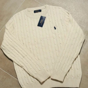 Vit kabelstickad tröja Polo Ralph Lauren - Snygg vit kabelstickad tröja från Polo Ralph Lauren med klassisk blå broderad logga på bröstet. Rund halsringning och ribbade muddar vid ärmslut och nederkant. Perfekt för dig som gillar stilrena och tidlösa plagg.