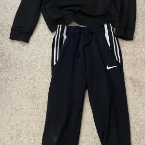 Svarta mjukisbyxor från Nike och adidas hoddie - Svarta mjukisbyxor från Nike med vita ränder längs sidorna och klassisk Nike-logga på benet. Byxorna har snörning i midjan och är gjorda i mjukt material som är perfekt för chill eller träning. Snygg och sportig look med normal passform. Adidas hoddie 11 åringar Nike byxor 128 till 137 cm