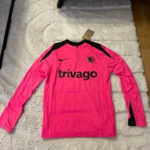 Chelsea långärmad rosa Nike fotbollströja - Ny med prislapp. Chelsea långärmad fotbollströja från Nike i stark rosa med svarta detaljer och logga. Tröjan har halv dragkedja framtill, svart lejonemblem på bröstet och svart 'trivago' tryck. Tillverkad i lätt, ventilerande syntetmaterial.