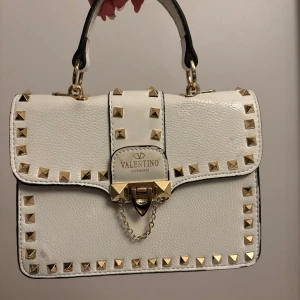 Vit Valentino handväska med nitar - Snygg vit handväska från Valentino Garavani i skinn med guldiga nitar runt kanterna och på locket. Väskan har både handtag och avtagbar axelrem, samt coolt lås med kedjedetalj. Insidan är röd och har fack för småsaker. Perfekt för dig som gillar statement-accessoarer.
