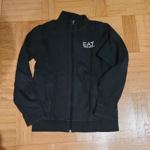 Svart trackjacka EA7 Emporio Armani - Svart trackjacka från EA7 Emporio Armani med hel dragkedja framtill och logga på bröstet. Jackan har hög krage, ribbade muddar och två fickor. Perfekt för en sportig och clean look.