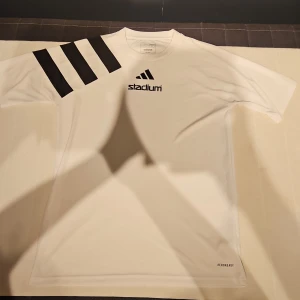 Vit Adidas t-shirt Stadium S - Vit t-shirt från Adidas med svarta detaljer och Stadium-tryck på bröstet. Klassiska tre svarta ränder på ena axeln. Regular fit och tillverkad i lätt polyester med AEROREADY-teknologi. Perfekt för sport eller chill.