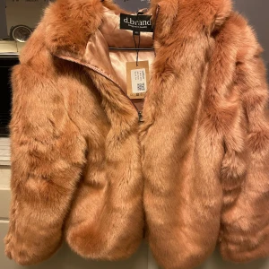 Rosa  pälsjacka med huva från d.brand - Supermjuk och fluffig rosa  pälsjacka från d.brand med huva och silkeslen insida. Jackan har dragkedja framtill och en riktigt cozy vibe, perfekt för kalla dagar. Snygg och trendig modell som ger en lyxig känsla.