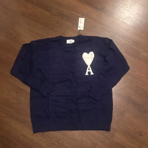 Mörkblå stickad tröja från Ami storlek M - Helt ny mörkblå stickad tröja från Ami Paris med vit hjärtlogga och bokstaven A på bröstet. Tröjan har rund hals, ribbade muddar och är långärmad. Perfekt för dig som gillar stilrena detaljer och vill ha något som sticker ut.
