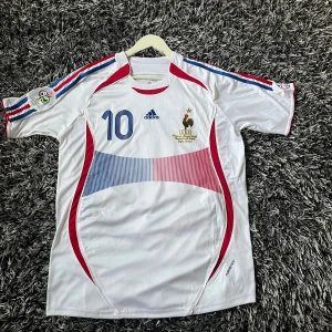 Frankrike Zidane Adidas fotbollströja - Säljer en klassisk vit fransk replika landslagströja från Adidas med nummer 10 och Zidane på ryggen. Tröjan har blå och röda detaljer, EM 2006 patch på ärmen och broderat landslagsemblem. Tillverkad i lätt polyester med korta ärmar och snygga ränder på axlarna. En perfekt tröja för fotbollsfantaster eller samlare. Hör gärna av er vid frågor. Skick 9,5/10. Pris kan diskuteras.
