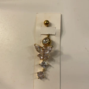 Guldig navelpiercing med fjärilsberlock från glitter.  - Snygg navelpiercing i guldton med glittrande stenar och en hängande fjärilsberlock. Stenarna är klara och ger ett lyxigt intryck. Perfekt accessoar för att lyfta din stil med lite extra bling.