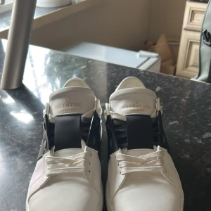 Valentino Garavani Open sneakers vit/svart - ( passar även 43)Säljer ett par Valentino Garavani Open sneakers i vitt skinn med svart band över mitten. Klassisk chunky sula och coola nitar baktill. Guldlogga på plösen och vita snören. Perfekta för dig som vill ha en lyxig och stilren sneaker med edge. PRIS KAN DISKUTERAS