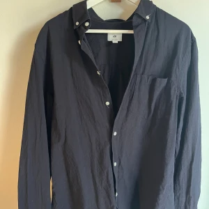 Svart skjorta från H&M i bomull - Svart långärmad skjorta från H&M i regular fit. Skjortan har klassisk krage med knappar, bröstficka och vita knappar framtill. Tillverkad i mjuk bomull, perfekt för en clean och stilren look.