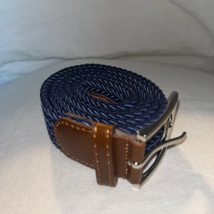 Mörkblå flätat bälte med skinn - Snyggt mörkblått flätat bälte med detaljer i brunt skinn och silvrigt spänne. Bältet har en stilren look med en twistad struktur och passar perfekt till jeans eller chinos. Skinnet ger en schysst kontrast mot det blåa materialet.