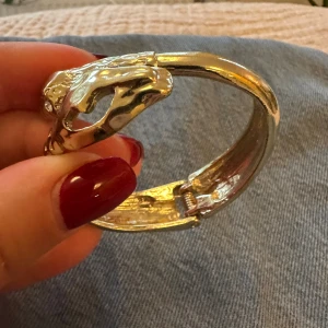 Guldigt armband med ormdetaljer - Unikt guldigt armband med två detaljerade ormar som möts framtill. Armbandet har en snygg, skulpterad yta med markerade fjäll. Perfekt statement piece för dig som gillar djurmotiv och coola accessoarer.