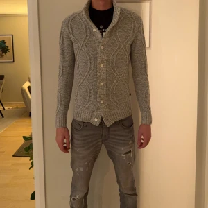 Grå stickad kofta från Soul Jeans - Säljer en grå stickad kofta från Soul Jeans med snygg kabelstickad design och stora vita knappar. Koftan har hög krage och långa ärmar, perfekt för lager-på-lager. Passformen är normal och materialet känns mjukt och skönt mot huden.