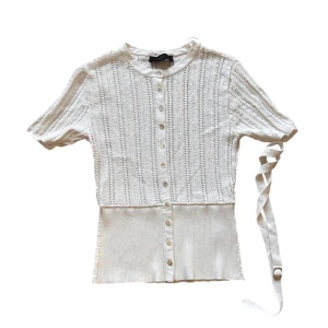 Vit stickad blus med bälte - ZARA - Supersnygg vit stickad blus från zara med korta ärmar och rund hals. Blusen har dekorativa knappar framtill och ett matchande bälte i midjan. Den är ribbad nedtill och har ett diskret hålmönster som ger en fräsch och modern vibe. Perfekt till jeans eller kjol!