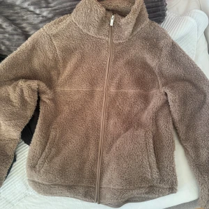 Beige teddyjacka med dragkedja - Mysig beige teddyjacka med hel dragkedja framtill och hög krage. Jackan är fluffig och har två fickor på sidorna. Perfekt för kyliga dagar och ger en avslappnad, trendig look.