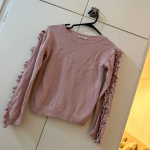 Rosa stickad tröja med volangärmar - Supersöt ljusrosa stickad tröja med volangdetaljer längs ärmarna. Tröjan har rund halsringning och ribbade muddar vid ärmslut och nederkant. Perfekt för dig som vill ha en gullig och trendig look.