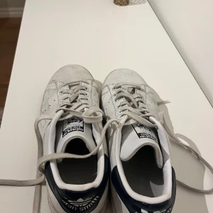 Adidas Stan Smith vita sneakers strl 42 - Mkt bra skick! Klassiska Adidas Stan Smith sneakers i vitt skinn med mörkblå detalj på hälen och perforerade ränder på sidorna. Skorna har snörning och platt sula, perfekt för en clean och tidlös look. Ikonisk logga på plösen och hälen.