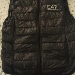 Svart dunväst från Emporio Armani EA7 - Stilren svart dunväst från Emporio Armani EA7 med quiltad design och dragkedja framtill. Västen har en diskret logga på bröstet och är tillverkad i lätt polyester som ger en cool look. Perfekt att slänga över en hoodie för extra värme.