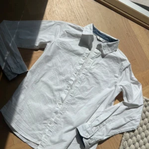 Vit skjorta från C, stl 140 - Klassisk vit skjorta från Cubus i storlek 140. Skjortan har lång ärm, knappar hela vägen framtill och en snygg krage med blå insida. Perfekt för dig som gillar stilrena och enkla plagg. Tillverkad i bomull för en skön känsla.