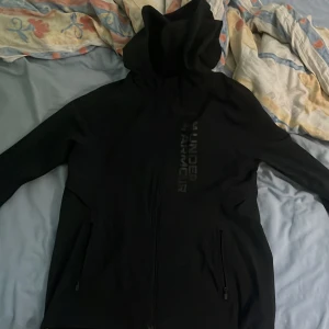 Svart vindjacka från Under Armour - Svart vindjacka från Under Armour med huva och dragkedja framtill. Jackan har diskret logga på bröstet, två fickor med dragkedja och en sportig, clean look. Perfekt för dig som gillar stilren och funktionell streetwear.