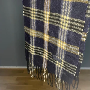 Burberry Halsduk Mörkblå - Snygg halsduk från Burberrys i klassiskt rutigt mönster med mörkblå, vita, svarta och röda detaljer. Gjord i 100% lammull och har fransar i ändarna. Perfekt accessoar för att lyfta din outfit med lite brittisk stil.
